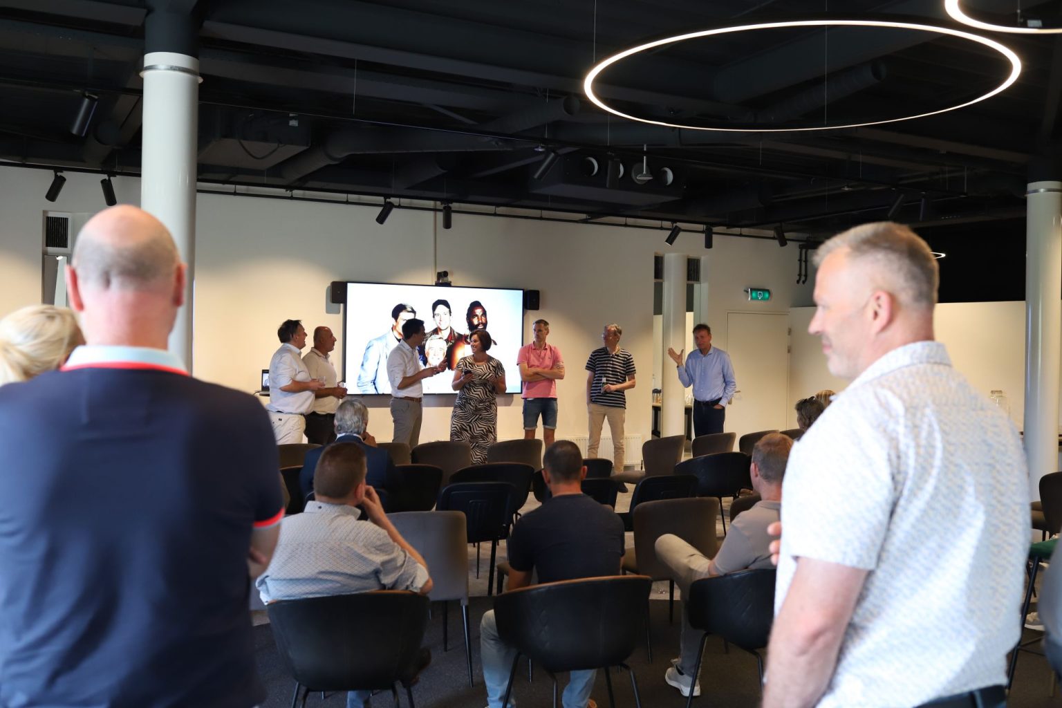 Een groep mensen in een moderne conferentieruimte kijkt naar een presentatie op een groot scherm met sprekers vooraan.