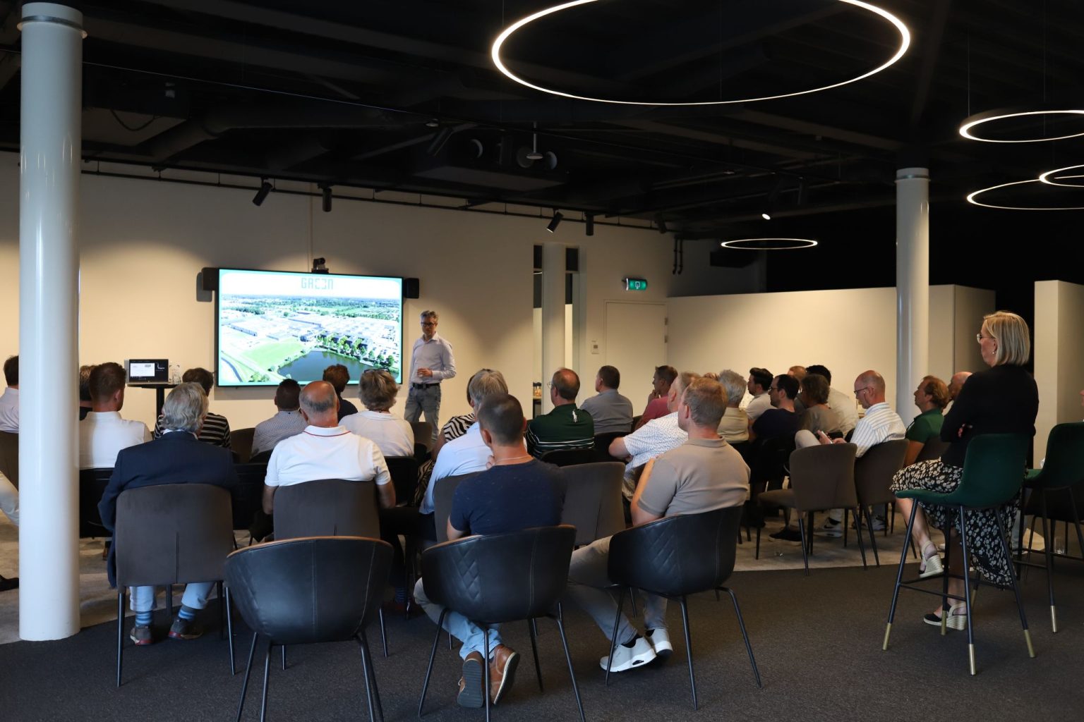 Een spreker presenteert aan een publiek in een moderne conferentieruimte met grote schermen en stijlvolle verlichting.