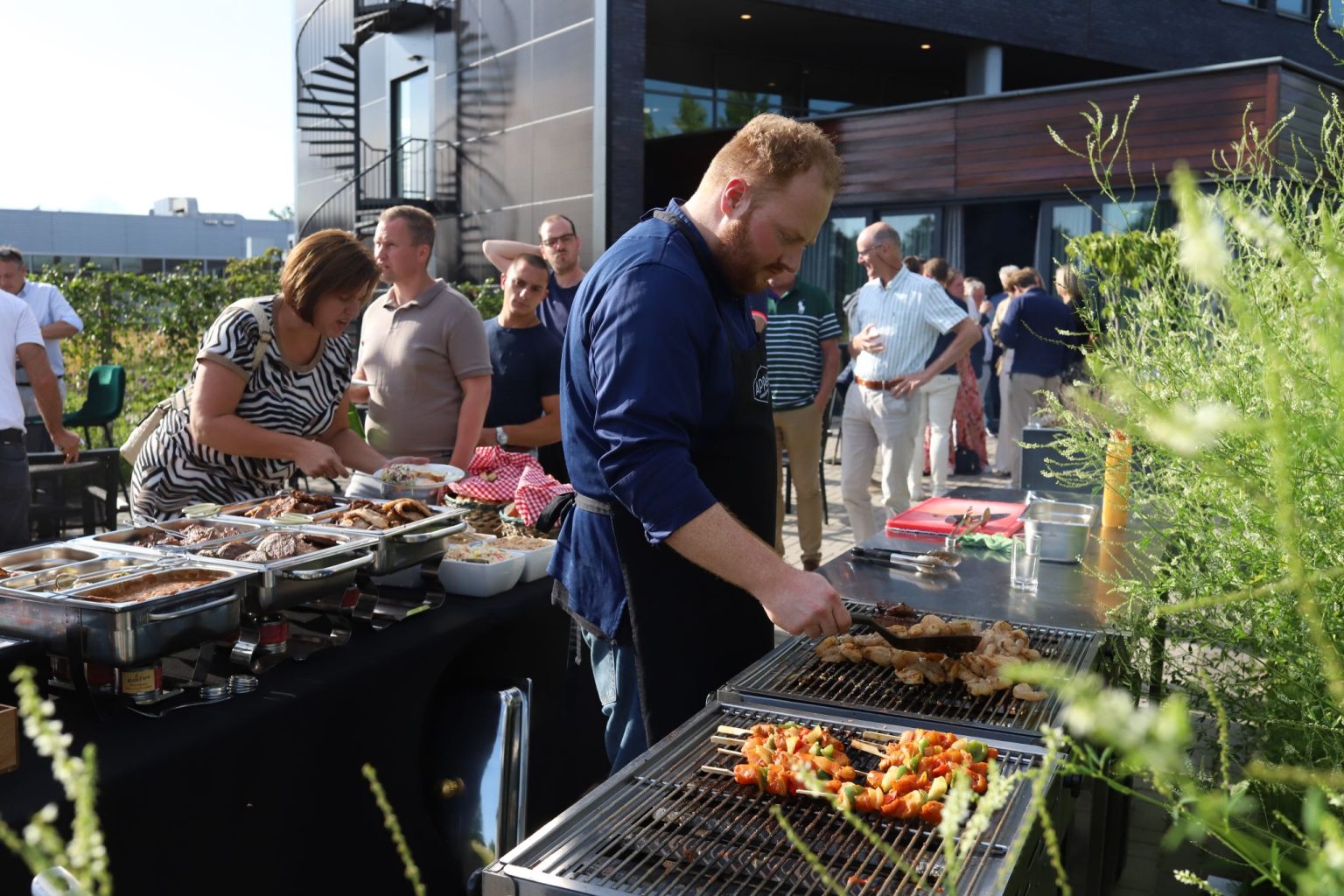 Man grilt vlees en groenten op barbecue tijdens buitenfeest met gasten en moderne architectuur op achtergrond