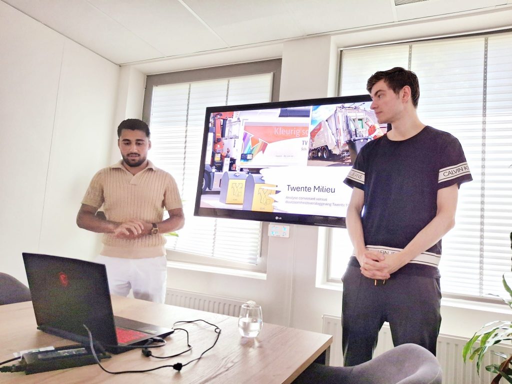 Twee mannen geven een presentatie in een kantoorruimte met een groot scherm en laptop op tafel.