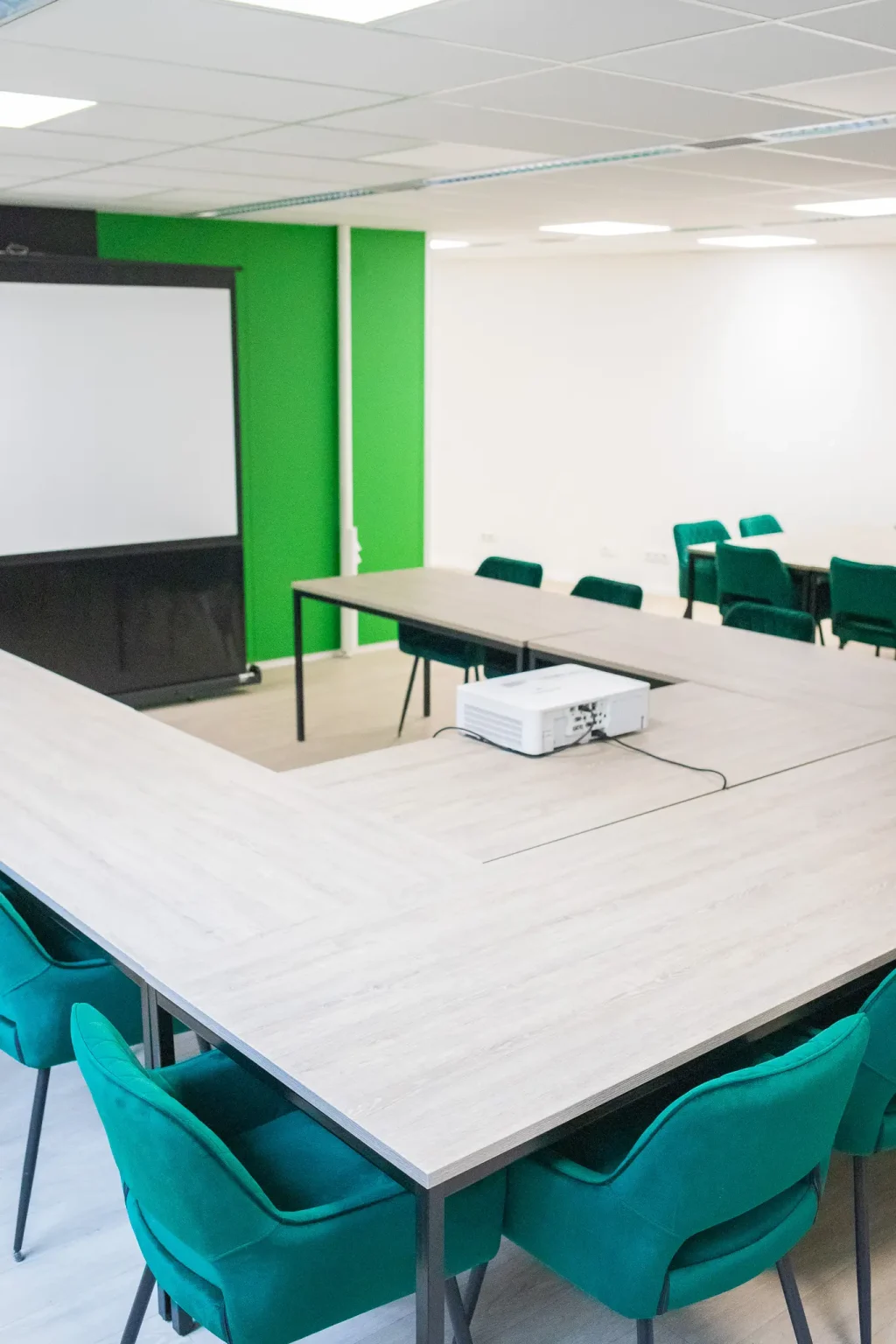 Moderne vergaderruimte met groene stoelen, witte tafels, projectiescherm en groene accentmuur in kantooromgeving