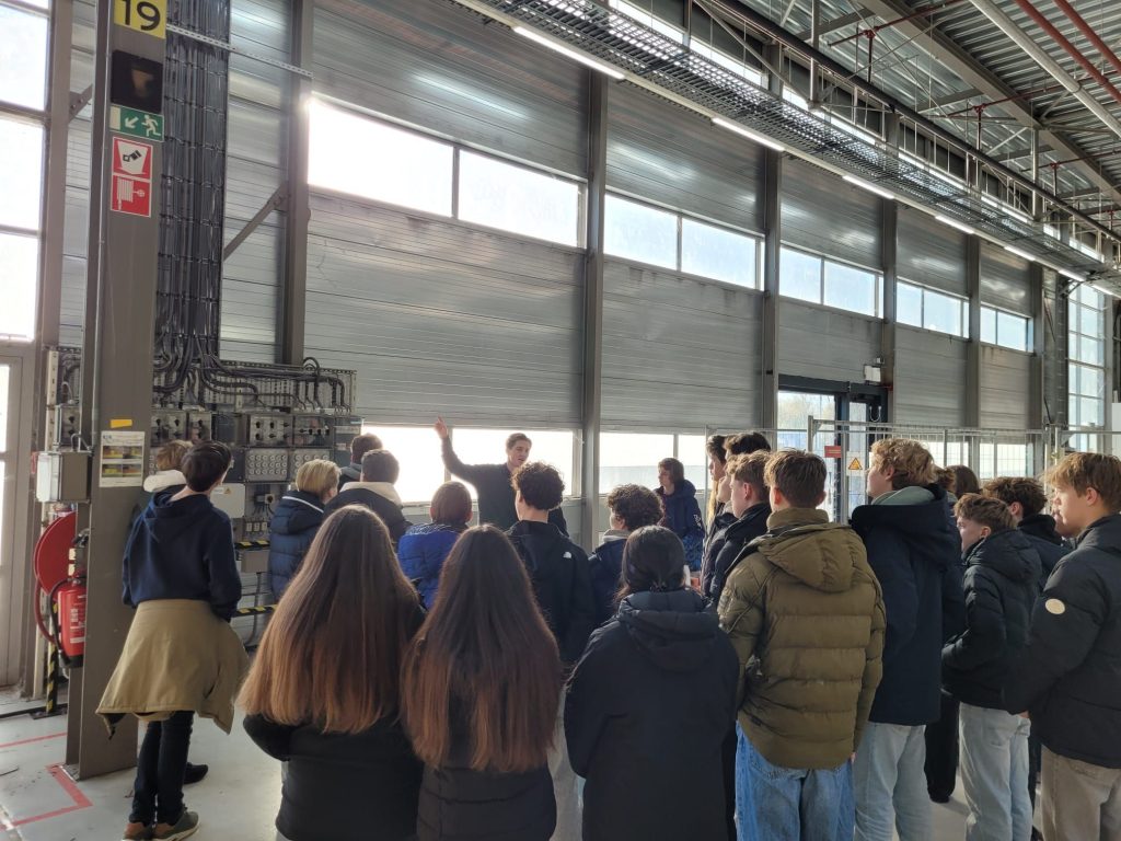 Groep studenten krijgt rondleiding in industriële fabriek met machines en transportband tijdens educatief bezoek