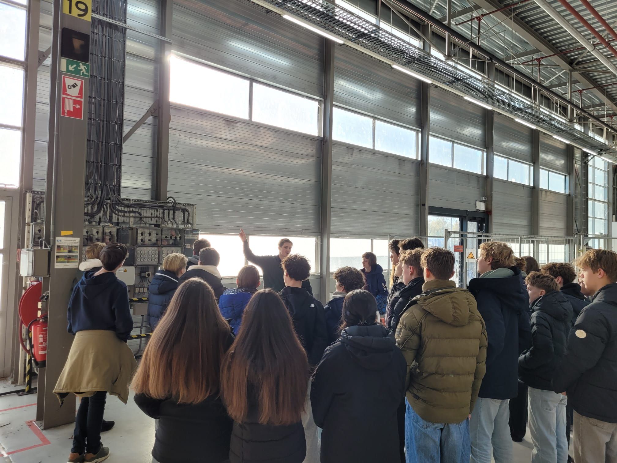 Groep studenten krijgt rondleiding in industriële fabriek met machines en transportband tijdens educatief bezoek