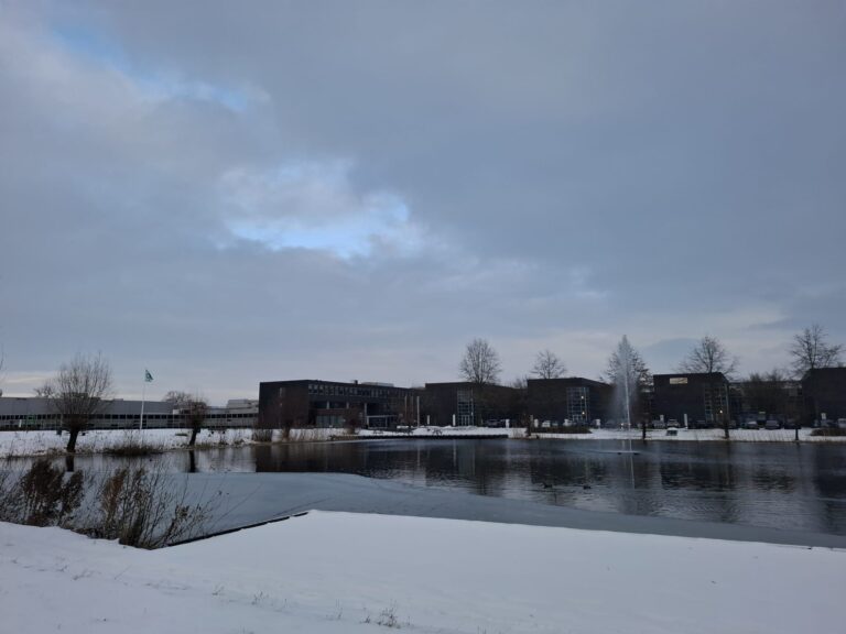 Winterlandschap met sneeuw, water en moderne gebouwen onder bewolkte hemel met kale bomen op de achtergrond