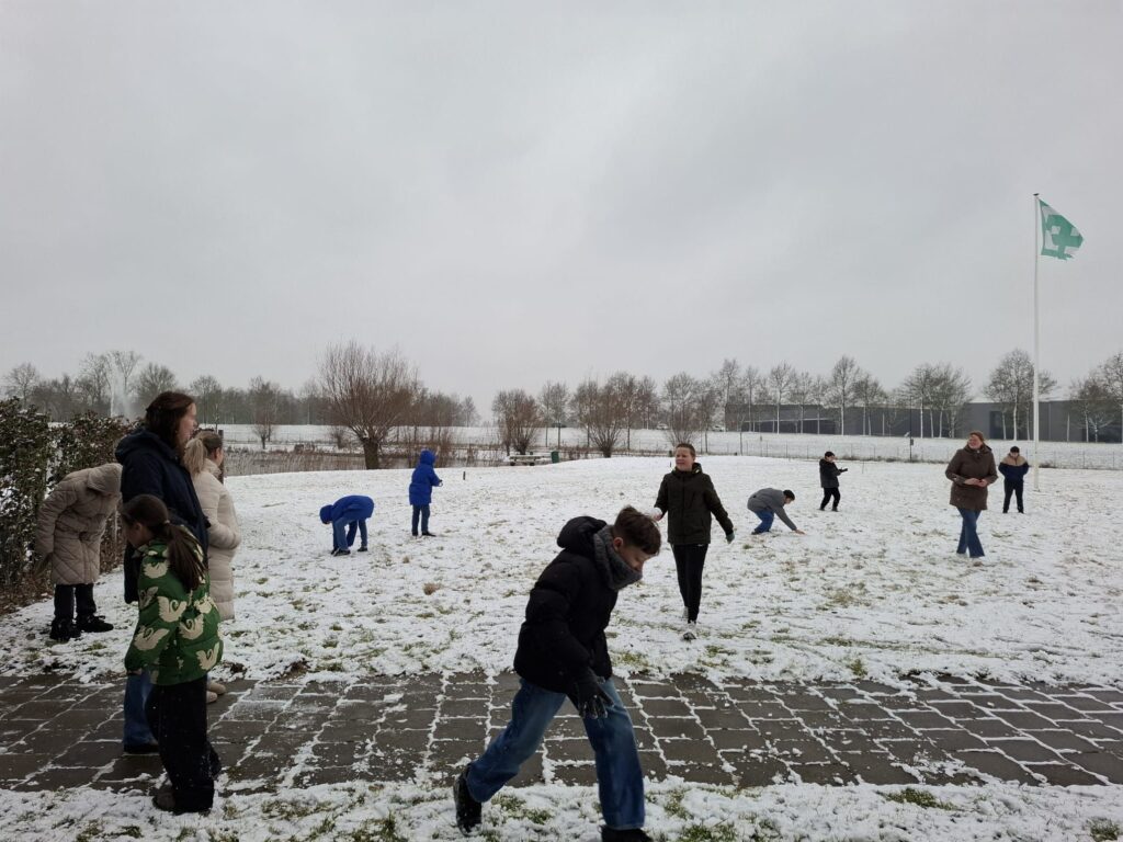 Mensen spelen in de sneeuw op een besneeuwd veld met op de achtergrond bomen, gebouwen en een Nederlandse vlag.