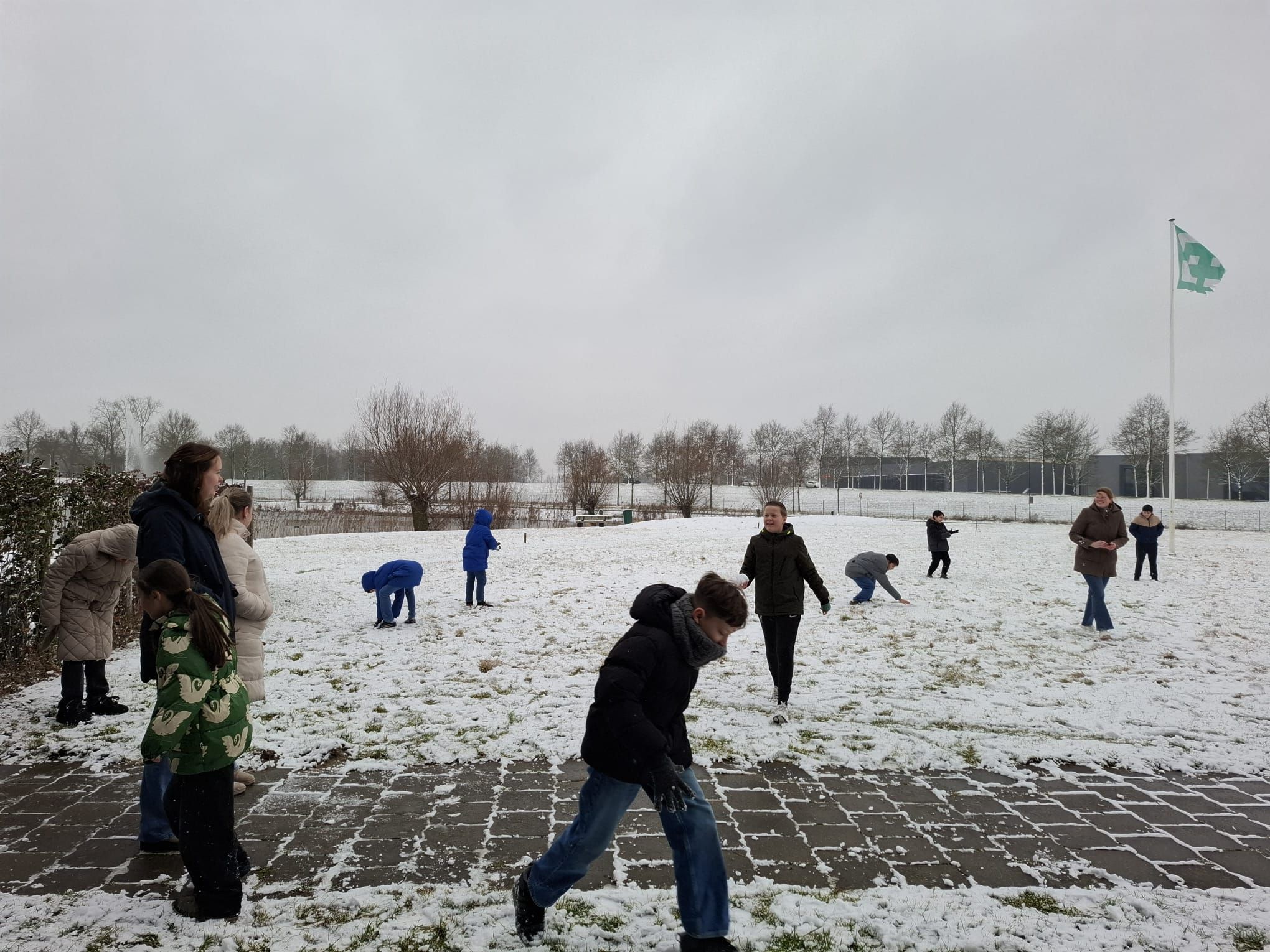 Mensen spelen in de sneeuw op een besneeuwd veld met op de achtergrond bomen, gebouwen en een Nederlandse vlag.