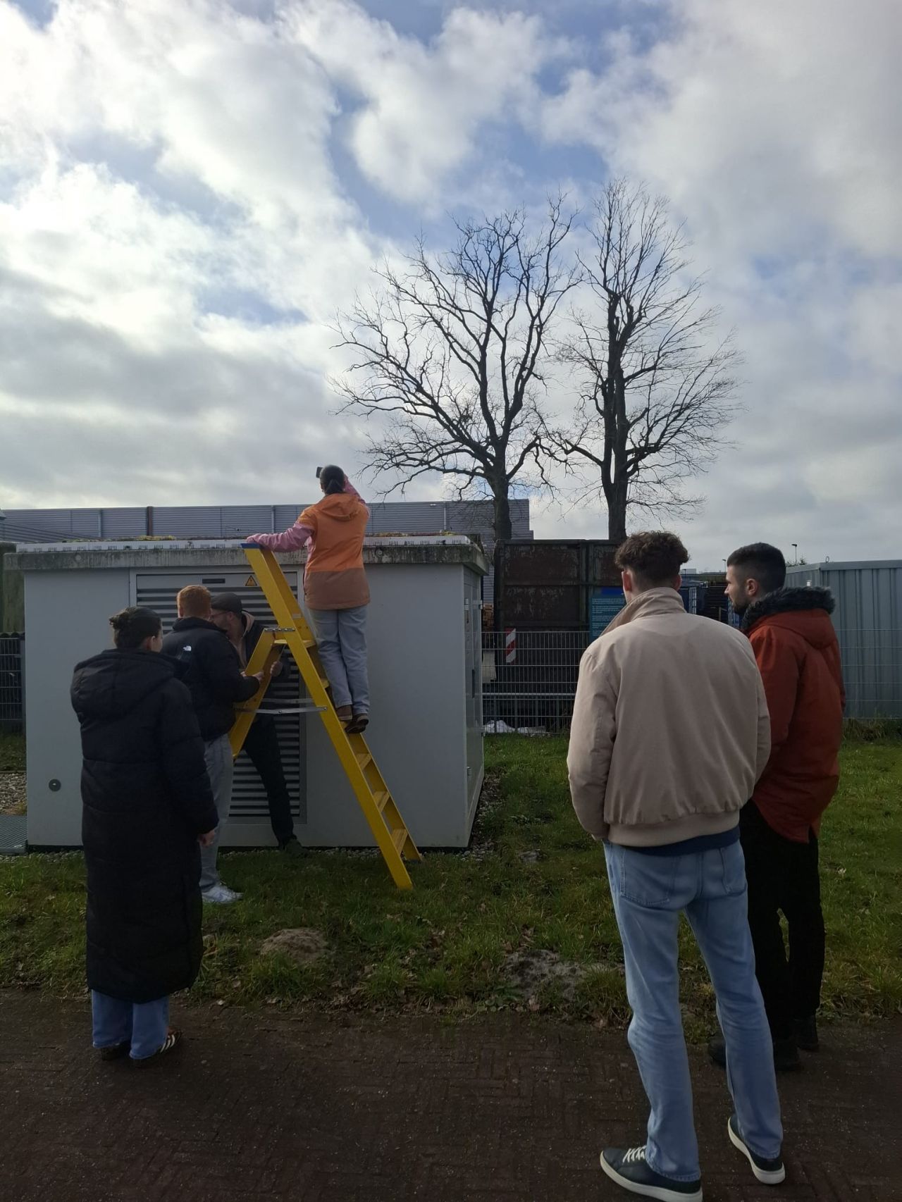Mensen installeren airconditioning unit met gele ladder terwijl anderen toekijken in buitenomgeving