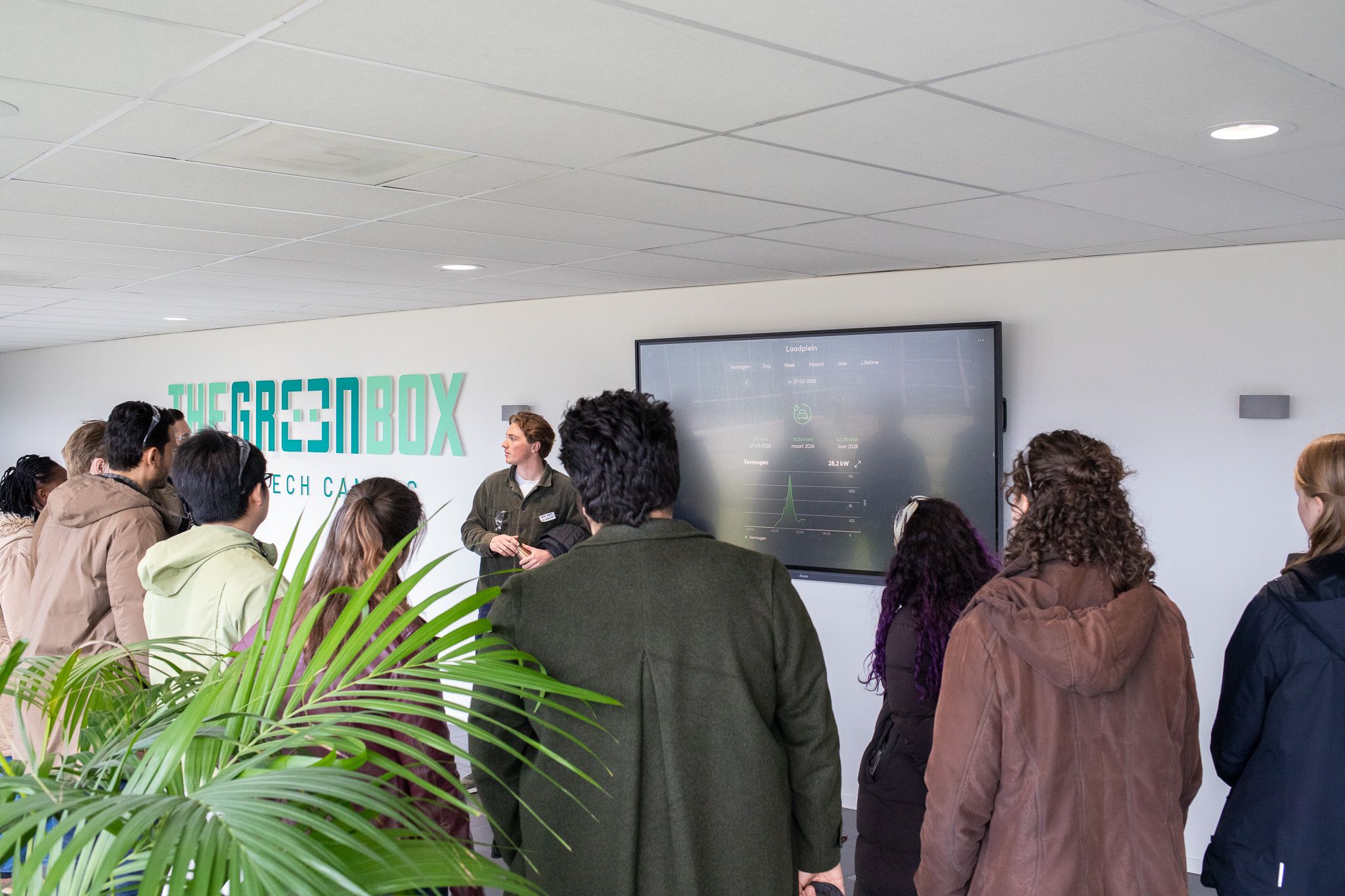 Een groep mensen luistert naar een presentatie over technologie bij The Green Box, met een groot scherm op de achtergrond.