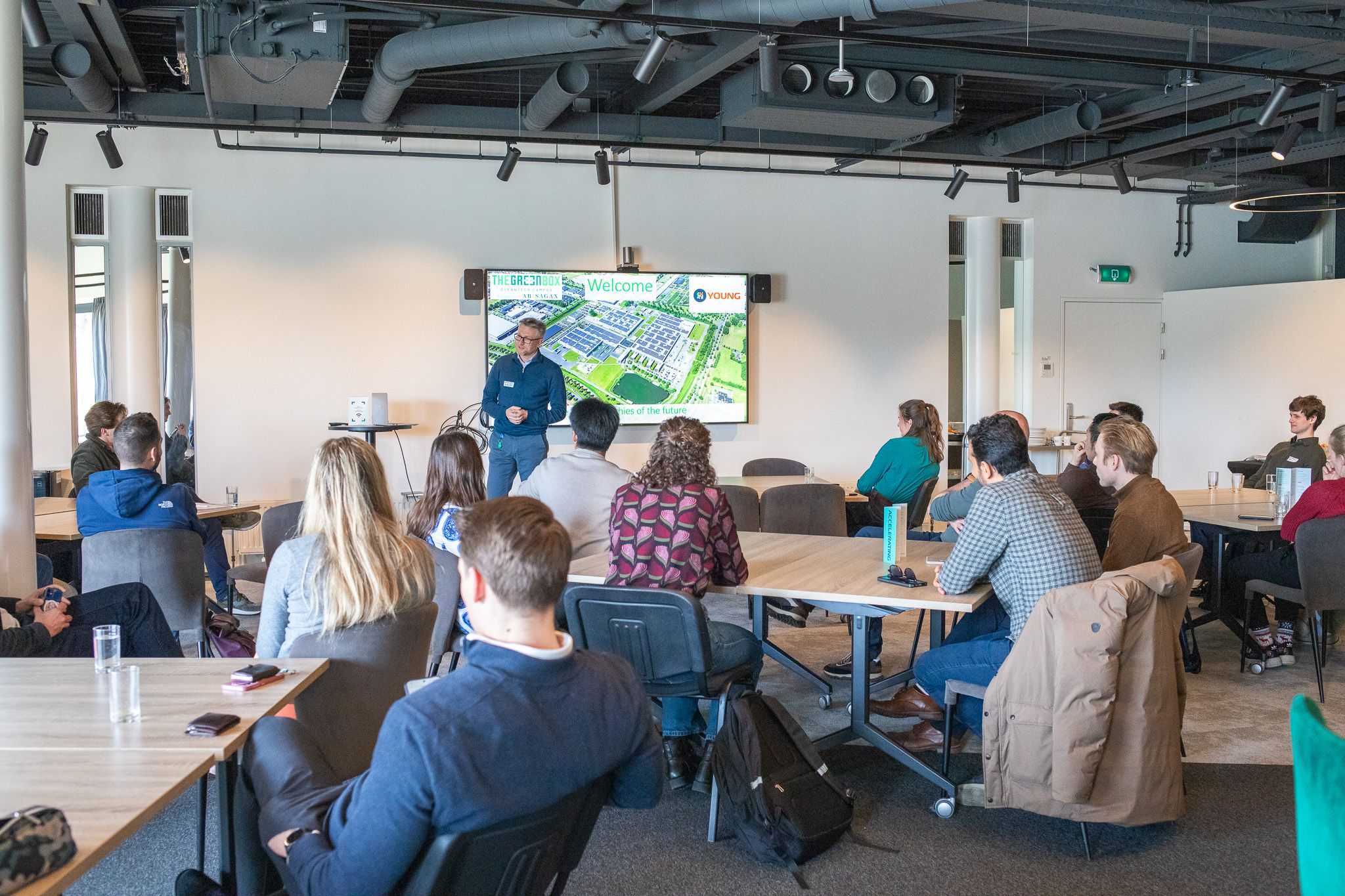 Een presentator geeft een lezing aan studenten in een moderne collegezaal met een groot scherm dat een campusplattegrond toont.