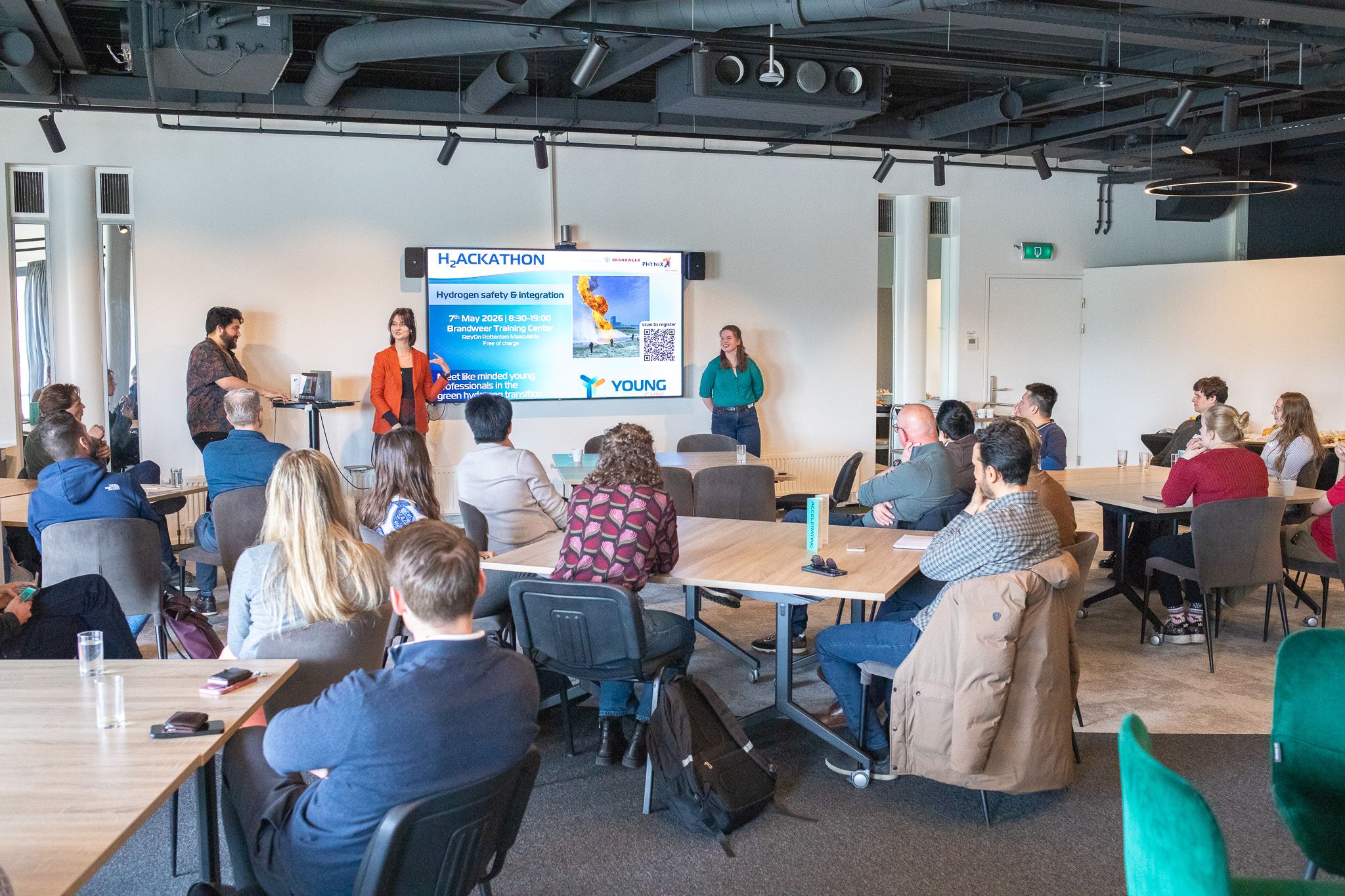 Presentatie over H2 Hackathon in moderne vergaderruimte met publiek aan tafels en sprekers bij groot scherm