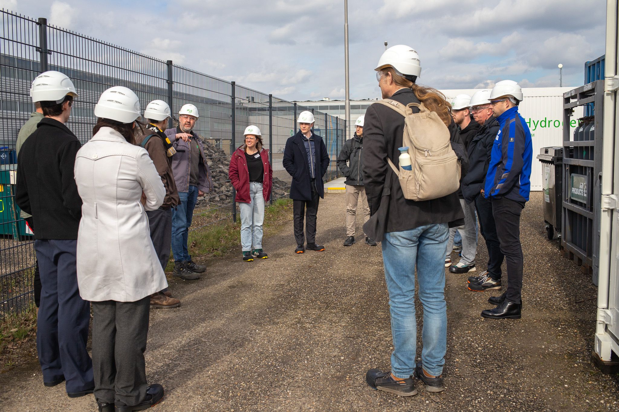 Groep mensen met witte helmen tijdens industriële rondleiding bij waterstoffaciliteit met hekwerk en gebouwen op achtergrond