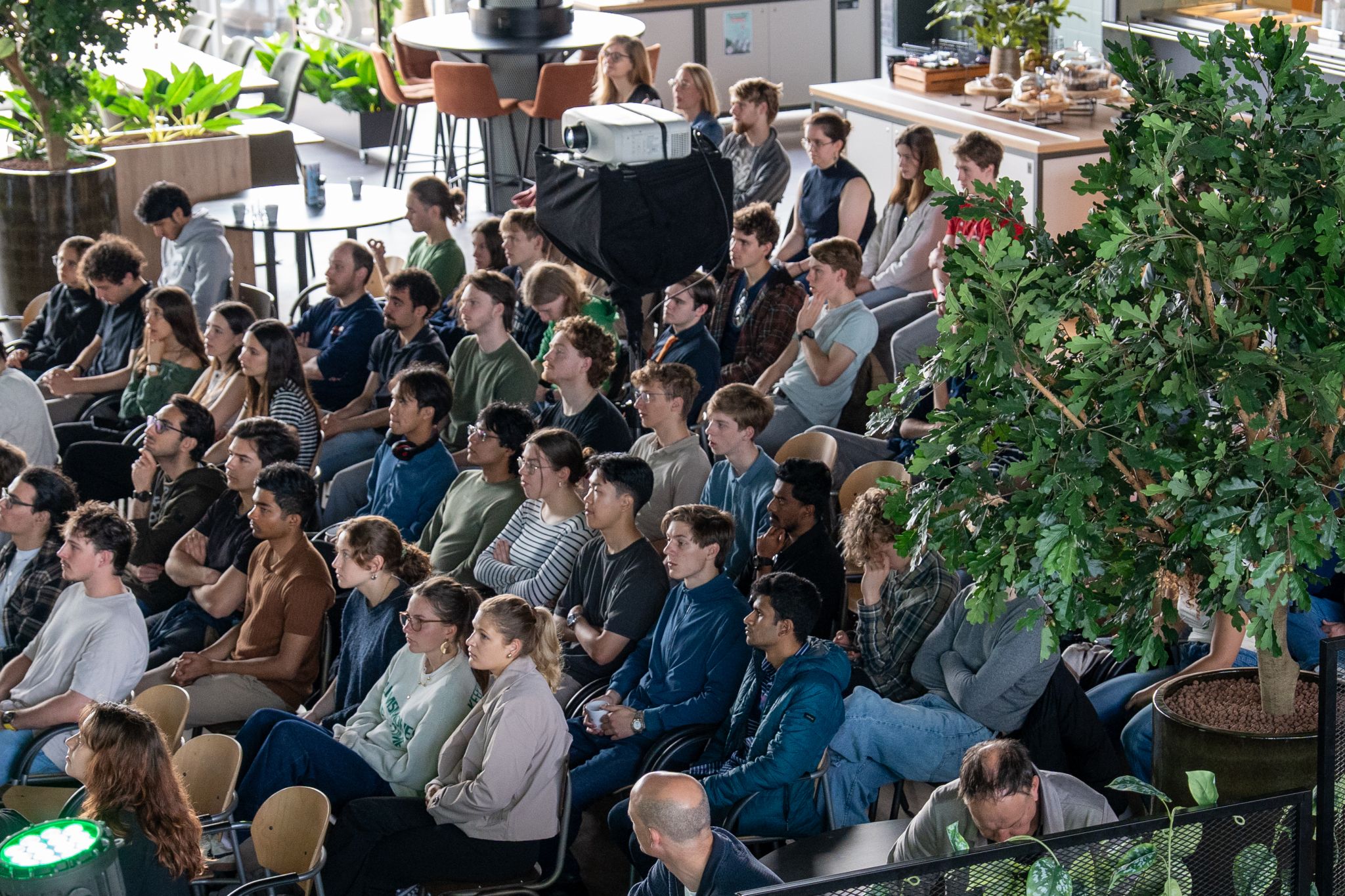 Een groep mensen zit in een moderne ruimte met veel planten en luistert naar een presentatie met een projector.