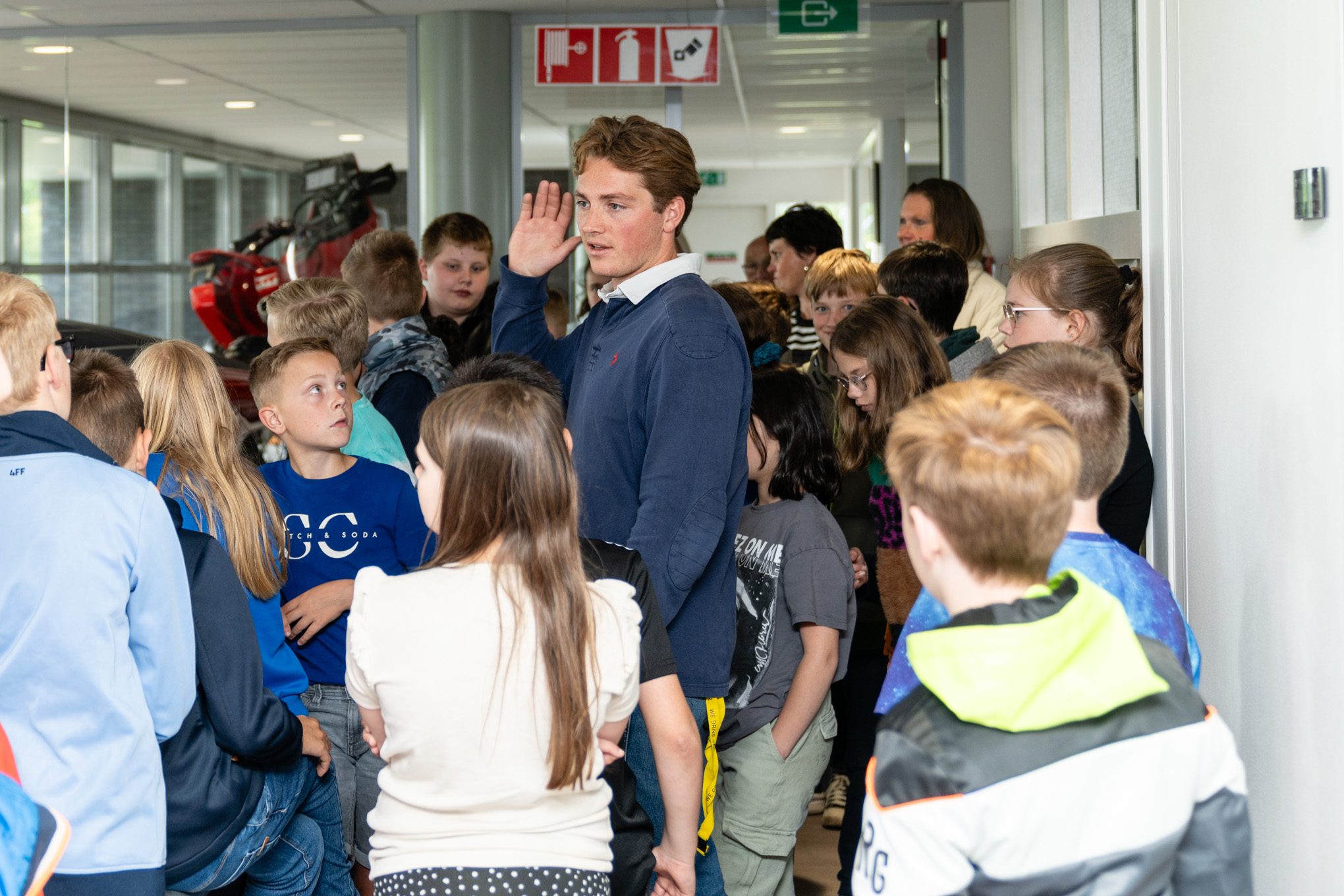 Jonge begeleider leidt groep kinderen door een gang tijdens een rondleiding in een bedrijfsgebouw.