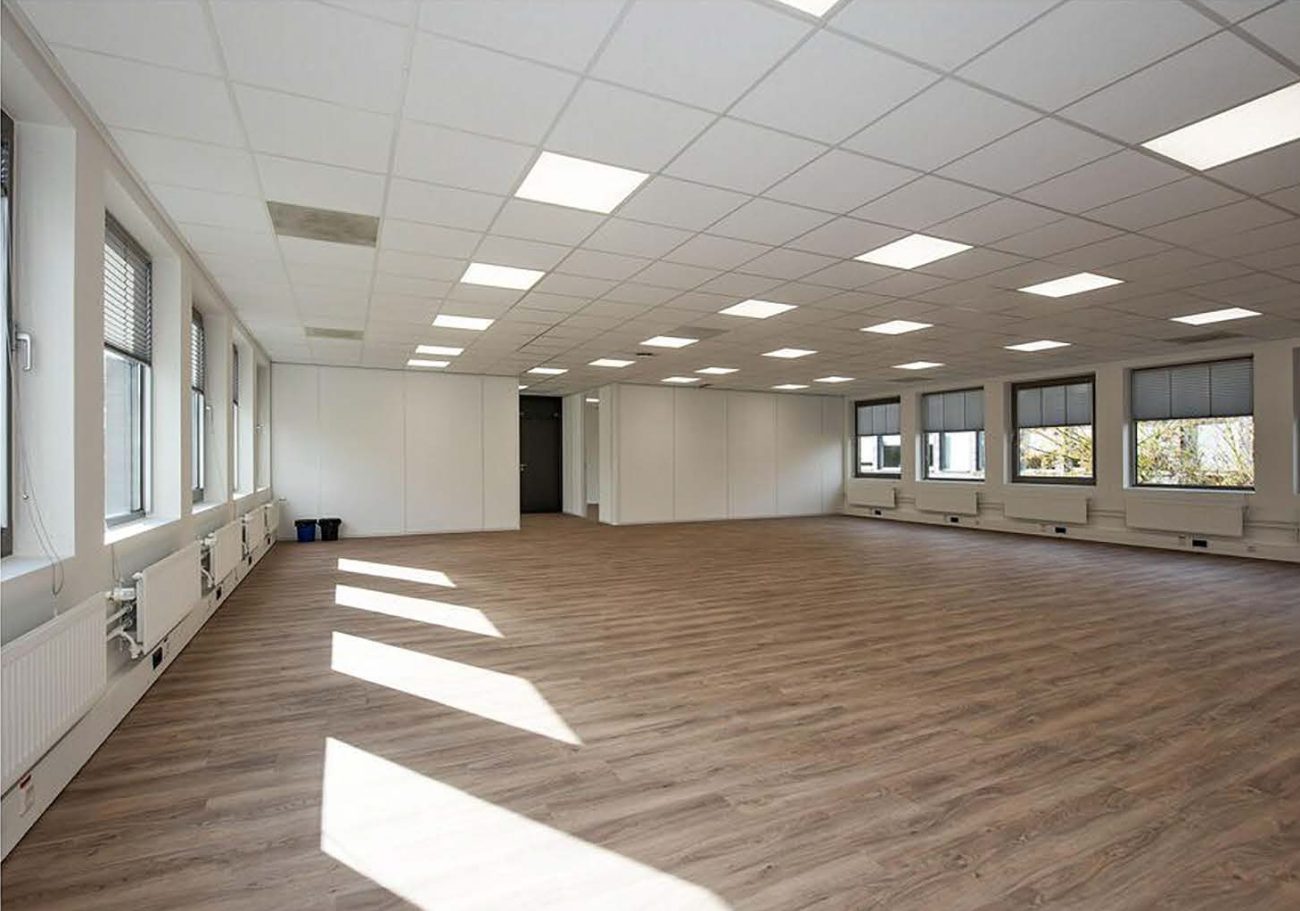 Lege kantoorruimte met houten vloer, witte wanden, verlaagd plafond en grote ramen met natuurlijk licht